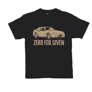 May include: Black t-shirt featuring a beige illustration of a classic car. The text "ZERO FOX GIVEN" is printed below the car graphic in a matching beige color. The shirt is made of a soft material.