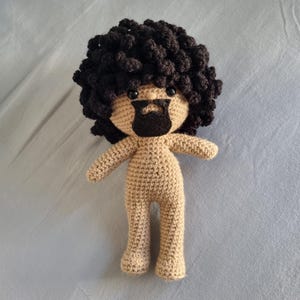 Può includere: Bambola all'uncinetto fatta a mano con corpo, braccia e gambe beige. La bambola ha capelli ricci marrone scuro e una barba nera. La bambola è in piedi e ha un design semplice e minimalista.