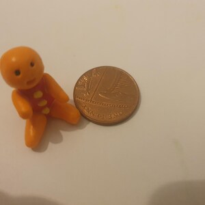 May include: A small, orange plastic gingerbread man figurine with a red vest and yellow buttons, next to a copper penny. The figurine is approximately 1 inch tall, a whimsical collectible.