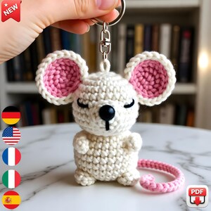 May include: A handmade crochet mouse keychain in white with pink accents. The mouse has large ears with pink centers and a pink tail. The keychain is held by a silver metal clasp. The word "NEW" is in a red banner in the top left corner.