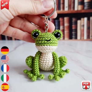 May include: A handmade crochet frog keychain in shades of green and cream. The frog has large black eyes and is attached to a silver keyring. The frog is a whimsical accessory.