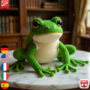 May include: A large, handmade crochet frog in shades of green and cream. The frog has large, expressive eyes and detailed features. The frog is sitting on a marble surface. A "NEW" tag is in the upper left corner.