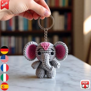 May include: Crocheted elephant keychain in gray with pink accents. The elephant has large, pink-lined ears and a pink heart on its head. The keychain is held by a hand, with a silver ring and chain. The background is a blurred bookshelf.