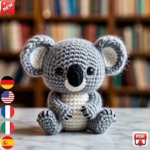 May include: A crocheted koala bear in grey and white, featuring black eyes and nose. The koala is depicted in a seated position. The background is blurred, showing a bookshelf. A red banner with "NEW" is in the top left corner.