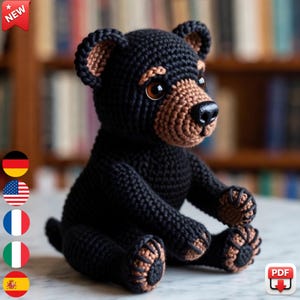Könnte beinhalten: Ein handgefertigtes Häkelhundespielzeug in Schwarz und Braun. Der Plüschhund hat braune Augen, eine braune Schnauze und braune Akzente an Ohren und Pfoten. Das Spielzeug sitzt auf einer weißen Oberfläche. Ein "NEU"-Aufkleber befindet sich oben links.