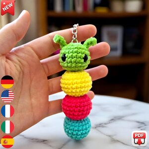 May include: A handmade crochet caterpillar keychain in green, yellow, red, and teal. The caterpillar has a silver keychain ring and black eyes. The word "NEW" is in a red banner in the top left corner.