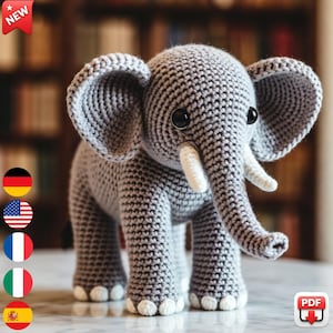 May include: A gray crocheted elephant with large ears, a long trunk, and white tusks. The handmade stuffed animal has black button eyes and is standing on a surface. A "NEW" tag is in the upper left corner.