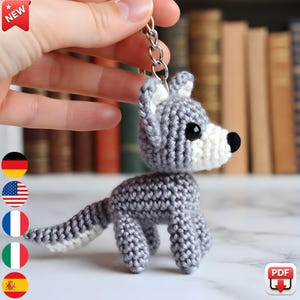 Könnte beinhalten: Ein handgehäkelter Wolf-Schlüsselanhänger in Grau und Weiß. Der Wolf hat schwarze Augen und eine schwarze Nase. Der Schlüsselanhänger ist an einem silbernen Metallring befestigt. Das Wort "NEW" steht in einem roten Banner oben links.