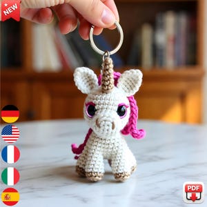 May include: A handmade white crochet unicorn keychain with a silver keyring. The unicorn has a brown horn, pink mane, and large, sparkly pink eyes. The keychain is held up by a hand.