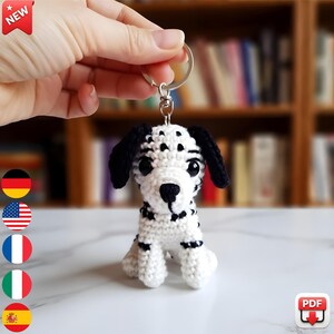 May include: A handmade crochet Dalmatian dog keychain. The small dog is white with black spots, ears, and a nose. The keychain is held by a silver ring. The background is blurred, with a bookshelf and a white surface.