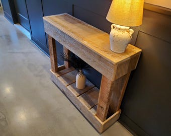 May include: A rustic wooden console table with a natural finish. The table features a lower shelf and is paired with a cream-colored lamp and a decorative vase. The table is made of reclaimed wood and has a distressed look.