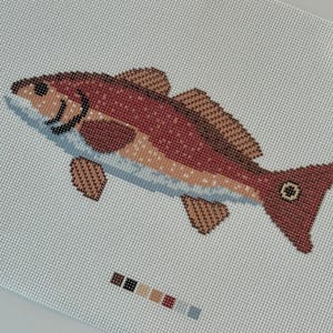 May include: A cross-stitch design of a fish in shades of red, brown, and blue. The fish is on a white background. A small color palette is below the fish.