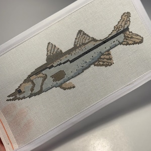 May include: A needlepoint canvas featuring a detailed depiction of a fish. The fish is rendered in shades of gray, brown, and beige, with a dark stripe along its side. The canvas is framed with a white border.