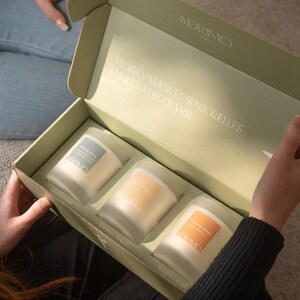 Peut inclure: Coffret cadeau ouvert contenant trois bougies parfumées. La boîte est vert clair avec le nom de la marque "ADRIMO" imprimé en haut. Les bougies sont dans des contenants en verre transparent avec des étiquettes. Le texte "TROIS EXPERIENCES SENSORIELLES UNIQUES À DÉCOUVRIR" est imprimé à l'intérieur.