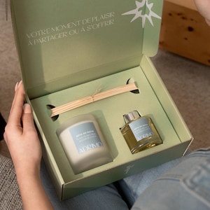 May include: An open gift box with a candle, diffuser, and reed sticks. The candle and diffuser have blue labels with text. The box is light green with text in French. The box is being held by a person.