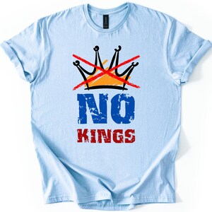 May include: Light blue t-shirt with a graphic design. The design features a black and orange crown crossed out with a red line, and the words "NO KINGS" in blue and red distressed font.