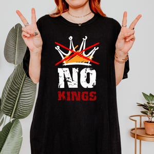 May include: Black t-shirt with a graphic design featuring a crossed-out crown and the words "NO KINGS" in white and red. The design includes orange and white accents.