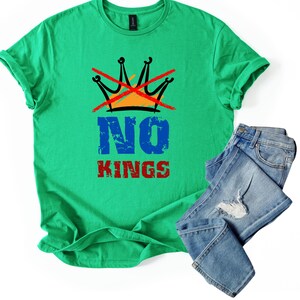 May include: A green t-shirt with the words "NO KINGS" in blue and red, and a crossed-out crown graphic. The shirt is paired with a pair of blue jeans.