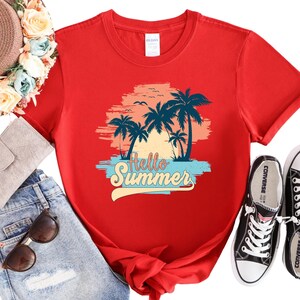 K&ouml;nnte beinhalten: Ein rotes T-Shirt mit einem tropischen Sonnenuntergangsdesign, Palmen und dem Text "Hello Summer". Die Grafik verwendet Blau-, Orange- und Wei&szlig;t&ouml;ne. Das Shirt wird mit einer blauen Jeans und schwarzen Converse-Sneakern kombiniert.