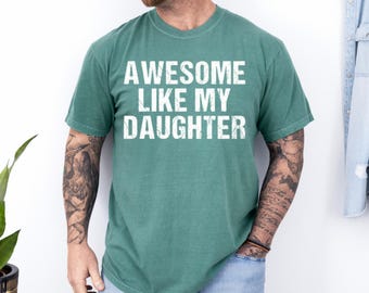 May include: A teal-colored t-shirt with the text "AWESOME LIKE MY DAUGHTER" in white block letters. The shirt is a solid color and has a crew neck. The image shows the shirt on a person.