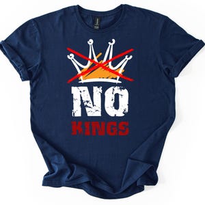 May include: Navy blue t-shirt with a graphic design. The design features a crown crossed out with a red line, the word "NO" in white, and the word "KINGS" in red. The shirt is folded.