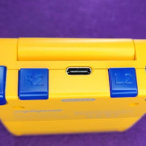 May include: A yellow handheld gaming device with blue accents. The device has blue buttons labeled "R", "R2", "L2", and "L". A USB-C port is visible. The device is set against a purple background.