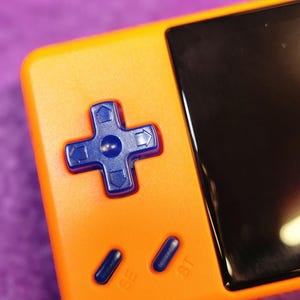 Op de afbeelding: Close-up van een oranje handheld gaming apparaat met een zwart scherm en een blauw kruis. Het apparaat heeft SE- en ST-knoppen. De achtergrond is een paars oppervlak.