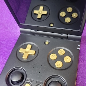 May include: Black handheld gaming console with gold-colored buttons, including a directional pad, action buttons, and a D-pad. The console is open, revealing a screen and additional controls. The SELECT and START buttons are visible.