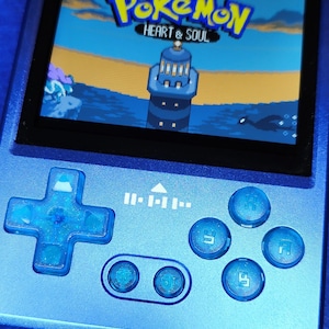 Pode incluir: Um console de jogos portátil azul exibindo o jogo "Pokémon Heart & Soul". O dispositivo possui um direcional, botões e uma tela mostrando uma cena do jogo com um farol. O dispositivo é azul.