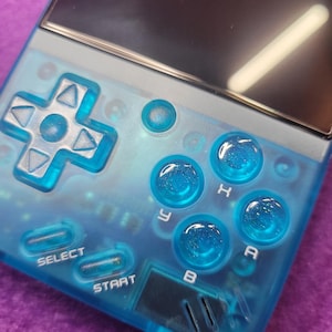 Könnte beinhalten: Eine transparente blaue Handheld-Spielkonsole mit einem Bildschirm und verschiedenen Tasten. Die Tasten umfassen ein Steuerkreuz, Select, Start und Aktionstasten mit den Bezeichnungen A, B, H und Y. Die Konsole hat ein schlankes, modernes Design.