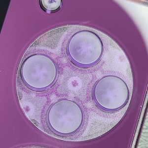 May include: Close-up of a purple handheld gaming device. The device features a circular control pad with four circular buttons and a directional button. The control pad has a silver and purple glitter design. The device is a vibrant purple color.
