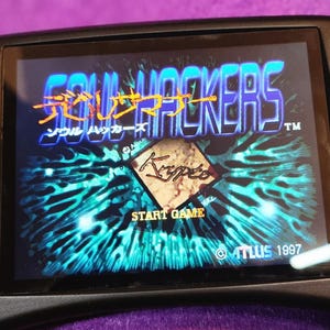 May include: A handheld video game console displaying the title screen of "Soul Hackers." The screen shows the game's logo in orange and blue, with Japanese text above the English title. The "Start Game" option is visible.
