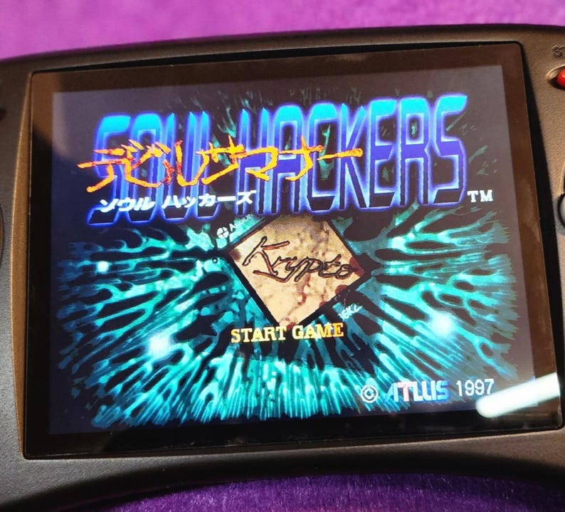 May include: A handheld video game console displaying the title screen of "Soul Hackers." The screen shows the game's logo in orange and blue, with Japanese text above the English title. The "Start Game" option is visible.