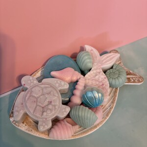 May include: A collection of pastel-colored soaps in the shape of sea creatures, including a turtle, seashells, and a mermaid tail, arranged on a decorative, leaf-shaped dish. The soaps are in shades of pink, blue, and white.