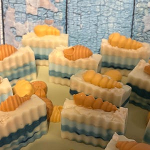May include: A collection of handmade soaps with a beach theme. The soaps are rectangular, with layers of white, blue, and cream colors. Each soap is topped with a small, orange seashell. The background is a light blue, wooden surface.
