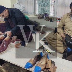 May include: Two men working on shoe construction. One is working on a pair of burgundy leather shoes with wooden shoe forms. The other man is working on a black leather item. Tools and materials are scattered around the workspace.