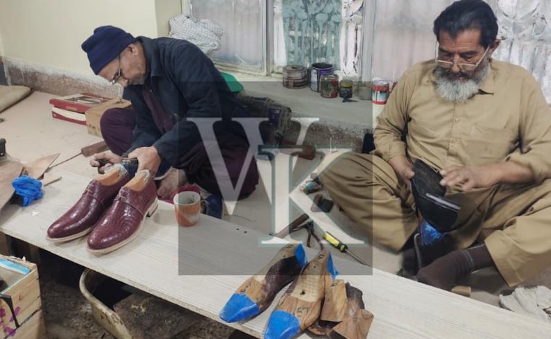 May include: Two men working on shoe construction. One is working on a pair of burgundy leather shoes with wooden shoe forms. The other man is working on a black leather item. Tools and materials are scattered around the workspace.