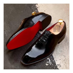 Peut inclure: Paire de chaussures de ville en cuir noir brillant avec semelles rouges. Les chaussures ont un design classique à lacets, un bout carré et un motif décoratif près de l'orteil. Des embauchoirs avec des boutons dorés sont à l'intérieur.