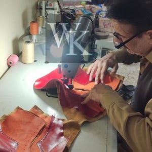 May include: A person using a sewing machine to assemble leather pieces. The leather is various shades of red and orange. Spools of thread and other sewing supplies are visible on the work surface. The image shows the process of creating leather goods.