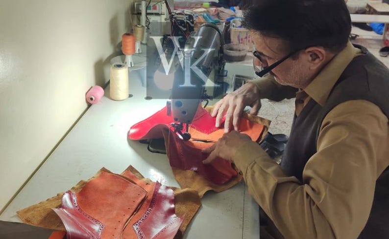 May include: A person using a sewing machine to assemble leather pieces. The leather is various shades of red and orange. Spools of thread and other sewing supplies are visible on the work surface. The image shows the process of creating leather goods.