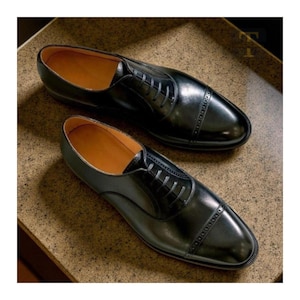 Puede incluir: Un par de zapatos de vestir negros de cuero, de estilo Oxford. Los zapatos tienen un acabado pulido, puntera y cierre de cordones. El interior es de color marrón claro.