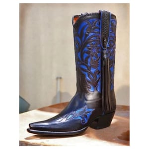 Könnte beinhalten: Ein schwarzer Leder-Cowboystiefel mit leuchtend blauer Blumenstickerei am Schaft und an der Schuhspitze. Der Stiefel verfügt über eine spitz zulaufende Form, einen Blockabsatz und eine lange, geflochtene Lederquaste an der seitlichen Zuglasche.