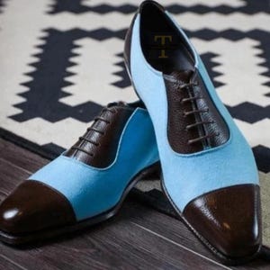 Op de afbeelding: Een paar tweekleurige geklede schoenen. De schoenen hebben een lichtblauw su&egrave;de bovenwerk met bruine lederen accenten op de neus, hiel en vetersluiting. De schoenen zijn geregen met bruine veters.