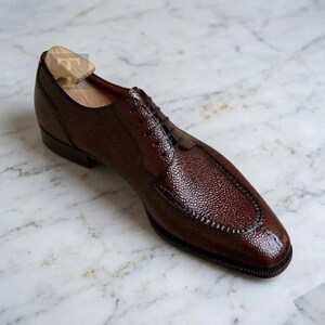 May include: A single brown leather dress shoe with a textured surface and a square toe. The shoe has a lace-up closure and a wooden shoe tree inside. The shoe is displayed on a marble surface.