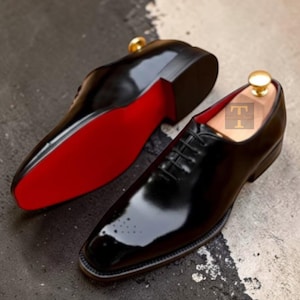 May include: A pair of black leather oxford shoes with a high-gloss finish, featuring a pointed toe with subtle brogue detailing, black laces, and a vibrant red sole. A wooden shoe tree with a gold-toned knob is inserted into each shoe.
