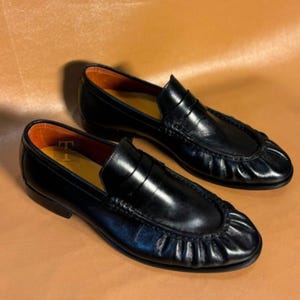 May include: A pair of black leather loafers with a classic design. The shoes have a rounded toe, a penny strap, and a stitched vamp. The interior is lined with a contrasting orange-brown material. The loafers are displayed on a brown surface.