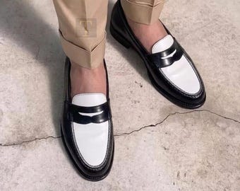 May include: Black and white leather penny loafers. The shoes have a classic design with a black upper, a white vamp, and a black strap across the top. The shoes are worn with tan pants.