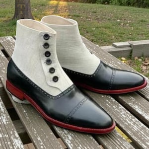 May include: A pair of two-tone ankle boots. The boots feature a black leather toe and heel, a cream-colored fabric upper, and a red sole. The boots have a button closure and are displayed on a wooden surface.
