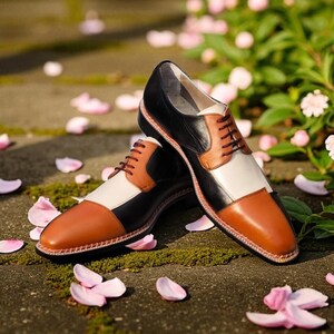 以下が含まれることがあります： ツートンカラーの革製ドレスシューズのペア。黒、白、茶色のパネルを使用したカラーブロックデザインが特徴です。靴は茶色の靴紐で、ピンクの花びらが散りばめられた石の表面に置かれています。