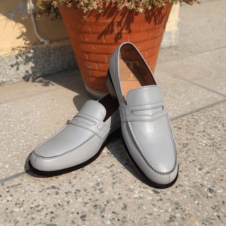 May include: A pair of light gray leather loafers with a classic design. The shoes have a rounded toe, a strap across the vamp, and a stitched sole. The loafers are displayed on a stone surface.